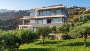 Una casa moderna de diversos pisos amb grans finestres, un balcó i una paret de pedra en una zona amb zones verdes i paisatge muntanyós.