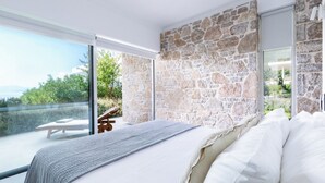Quarto com parede de pedra, uma cama grande e vista para o verde através de uma porta de correr de vidro.