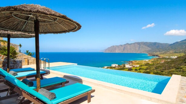 Ein Pool mit Blick auf das Meer und die Berge, Liegestühle und ein Reetdachschirm.