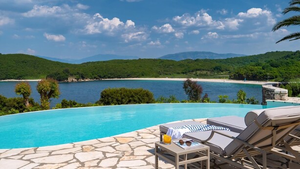 Un espace aménagé au bord de la piscine, avec des chaises longues et une table, offrant une vue sur une plage et des collines verdoyantes.