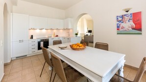 Une cuisine moderne avec des meubles blancs, un coin repas avec une table blanche et des chaises en osier, et un tableau accroché au mur.