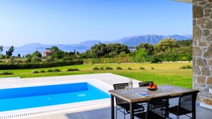 Un espace aménagé au bord de la piscine, avec une table et des chaises, offrant une vue sur la mer et les montagnes en arrière-plan.