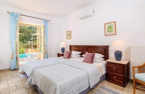 A bedroom with a double bed, two bedside tables, a chair, a window with curtains, and a view of a pool and greenery outside. - Cosy Villa with Sea-and-Mountain Views near Agios Nikitas (Lefkada)