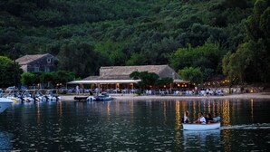 Ein See-Restaurant mit Außenbestuhlung, in der Nähe liegen Boote vor Anker, und ein bewaldeter Hügel im Hintergrund.
