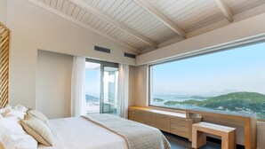 A bedroom with a large bed, a wooden bench, a view of the mountains, and a sliding door leading to a balcony. - Luxury Villa with Mesmerising Sea-and-Island Views and Large Private Pool (Perigiali)