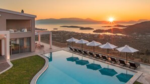 A modern hotel with a pool, sun loungers, and umbrellas, overlooking a scenic sunset view of a coastal area. - Luxury Villa with Mesmerising Sea-and-Island Views and Large Private Pool (Perigiali)
