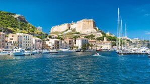 Un pueblo costero con un fuerte histórico en una colina, barcos amarrados a lo largo del malecón y edificios con balcones.