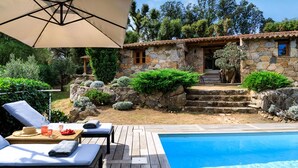 Zona junto a la piscina con sillones de descanso, una terraza de madera y un edificio de piedra con tejado de baldosas.