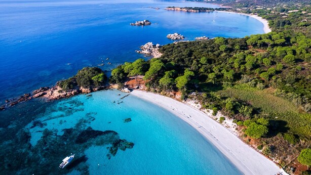 Vista aérea de una zona costera con playa arenosa, aguas azules cristalinas y vegetación verde exuberante.