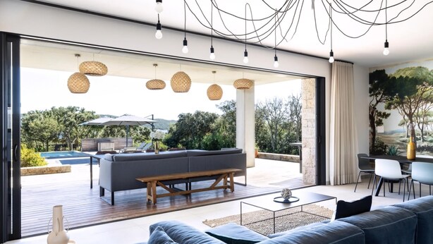 A modern living room with a large grey sofa, a wooden coffee table, and a view of an outdoor patio area with a pool and umbrellas. - Stylish Signature Villa with Heated Pool close to the Beach (Pianottoli-Caldarello)
