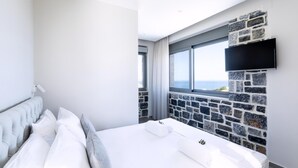 A hotel room with a large bed, a stone accent wall, a TV mounted above it, and a view of the sea through a window. - Mochlos Harbour View (Sitia)