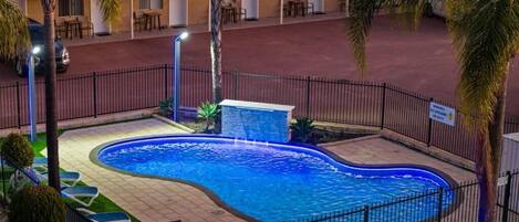 Uma piscina com borda curva, cercada por uma grade preta. Palmeiras e um edifício com telhado plano ao fundo.