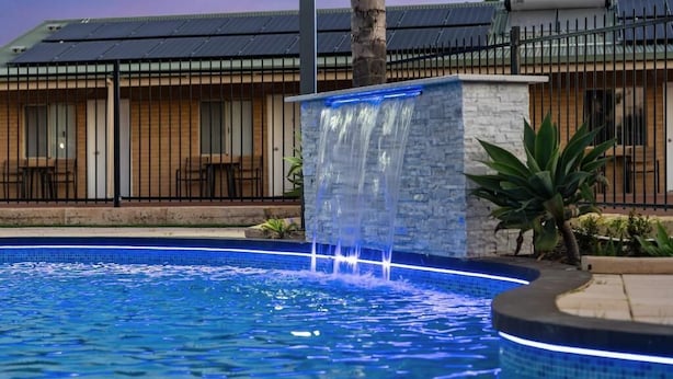 A swimming pool with a waterfall feature, surrounded by a stone wall and palm trees. - Pinjarra Resort (PINJARRA)
