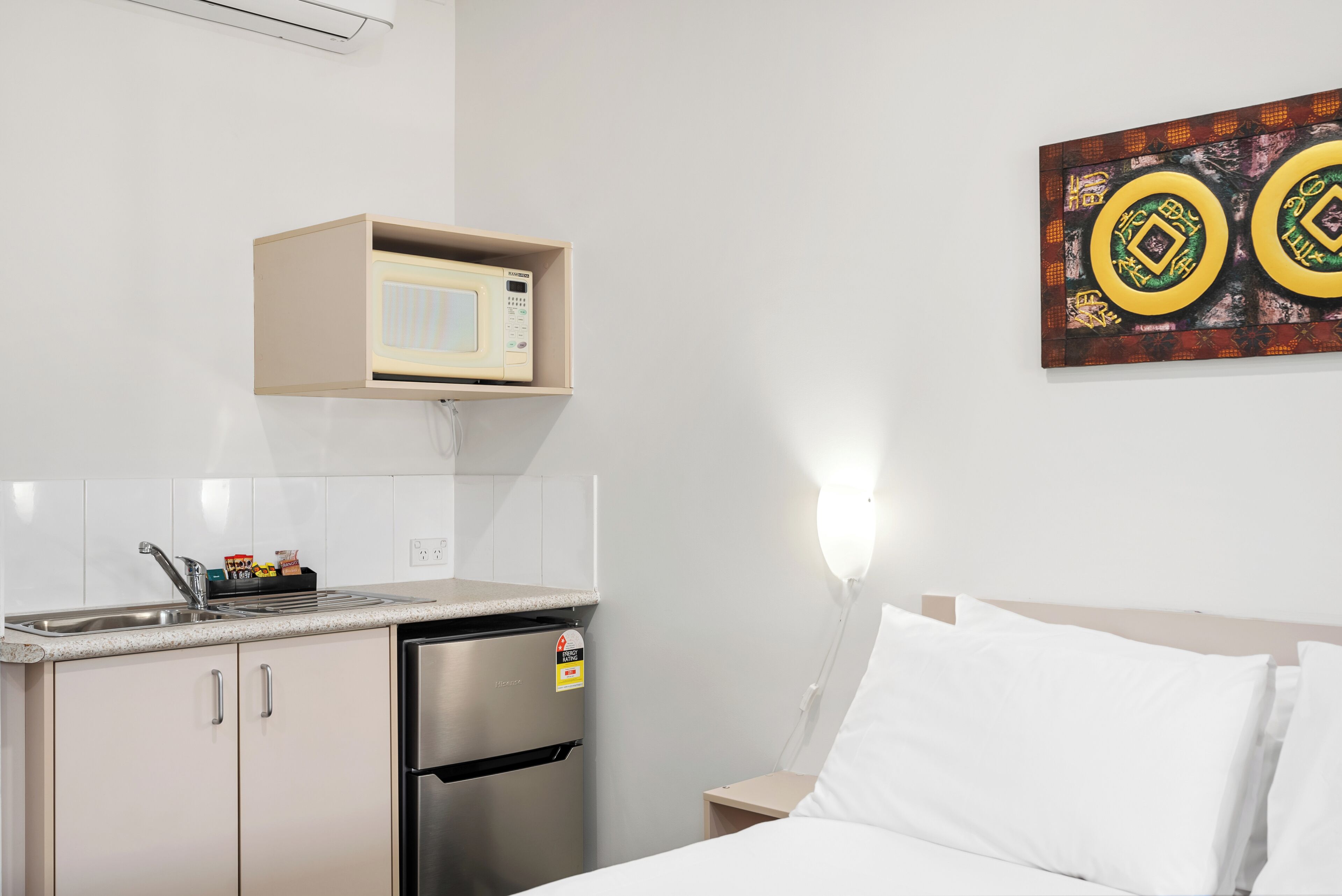 Une petite cuisine équipée d’un four à micro-ondes, d’un réfrigérateur et d’un évier. On y trouve un lit avec du linge de lit blanc et un luminaire fixé au mur.