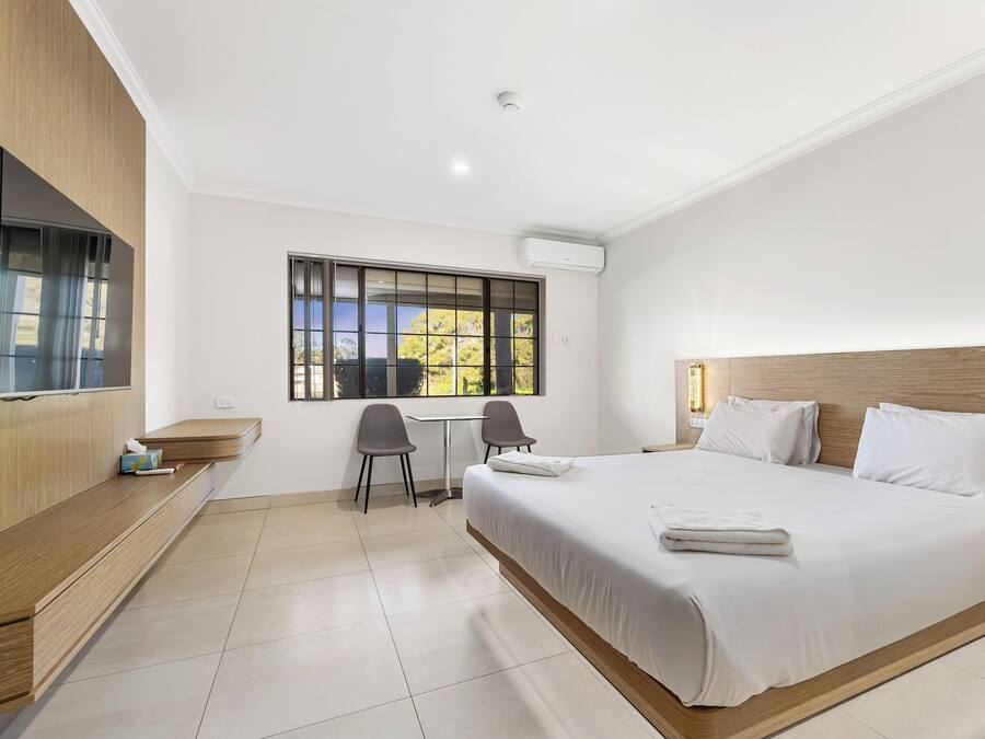 A hotel room with a large bed, a desk, two chairs, a TV, and a window with a view of trees.