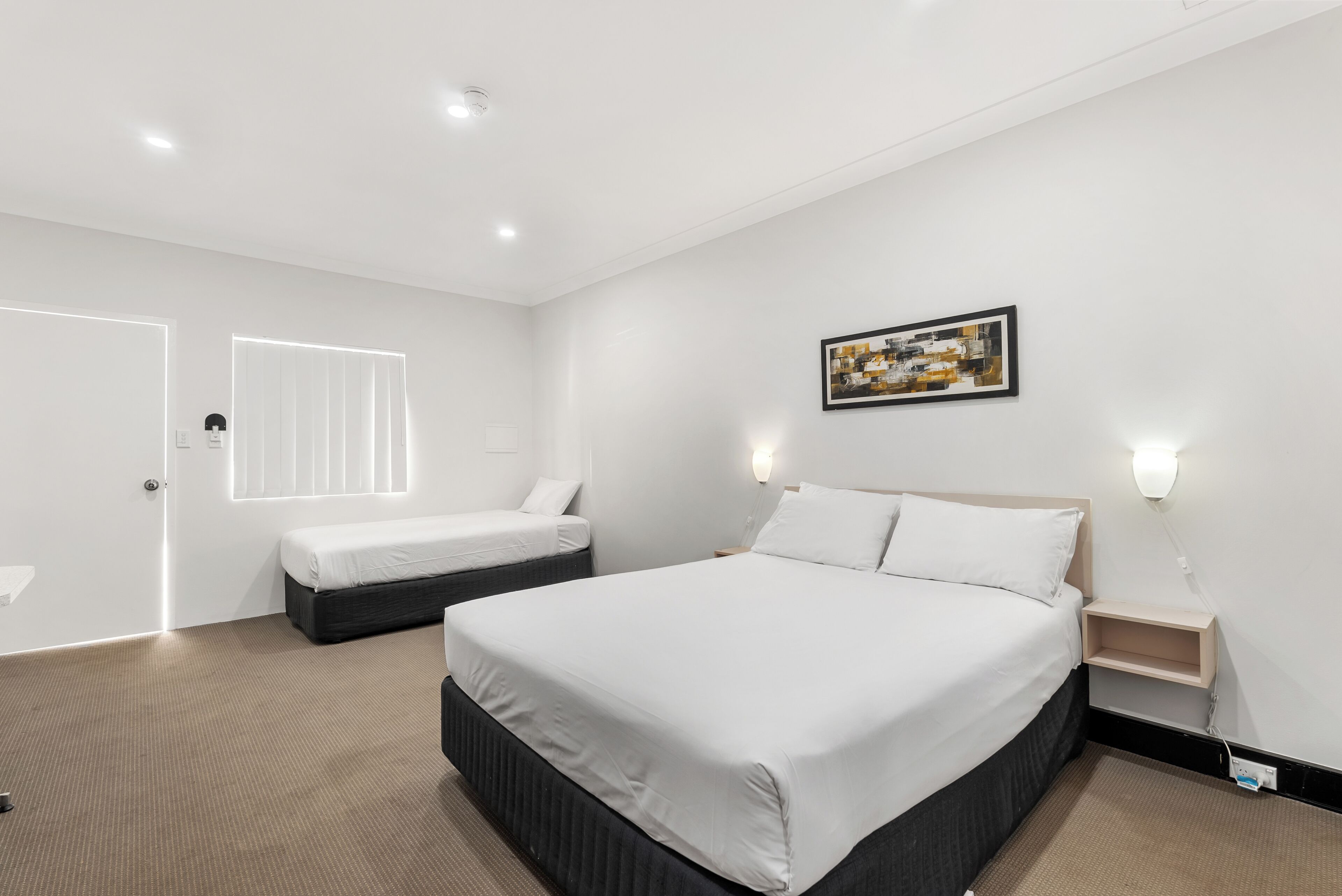 Quarto de hotel moderno com uma cama grande, uma cama menor, uma mesa de cabeceira, uma obra de arte na parede e uma janela com persianas.