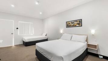Quarto de hotel moderno com uma cama grande, uma cama menor, uma mesa de cabeceira, uma obra de arte na parede e uma janela com persianas.
