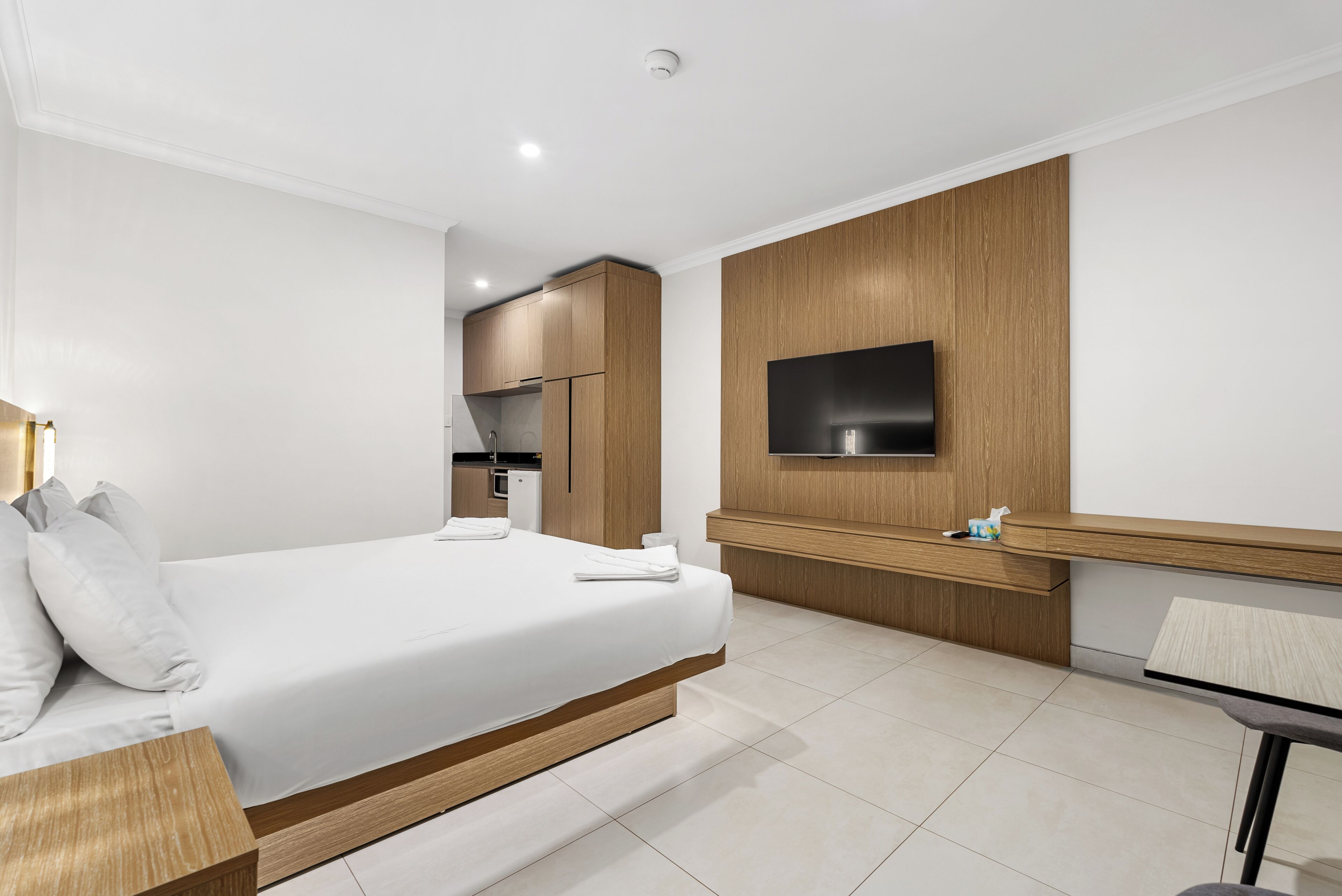 A modern hotel room with a bed, a desk, a flat-screen TV, and a kitchenette in the background.