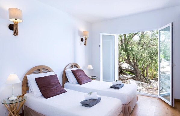 A modern hotel room with two beds, each with a wooden headboard, white bedding, and a view of a rocky landscape through a glass door.
