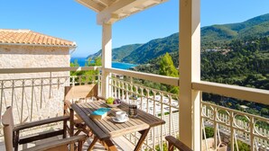 Un balcone con tavolo e sedie, vista su un paesaggio costiero con montagne e un cielo azzurro limpido.