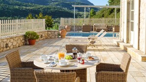 A sunlit outdoor dining area with wicker furniture, a pool, and a mountainous backdrop. - Cosy Villa with Sea-and-Mountain Views near Agios Nikitas (Lefkada)