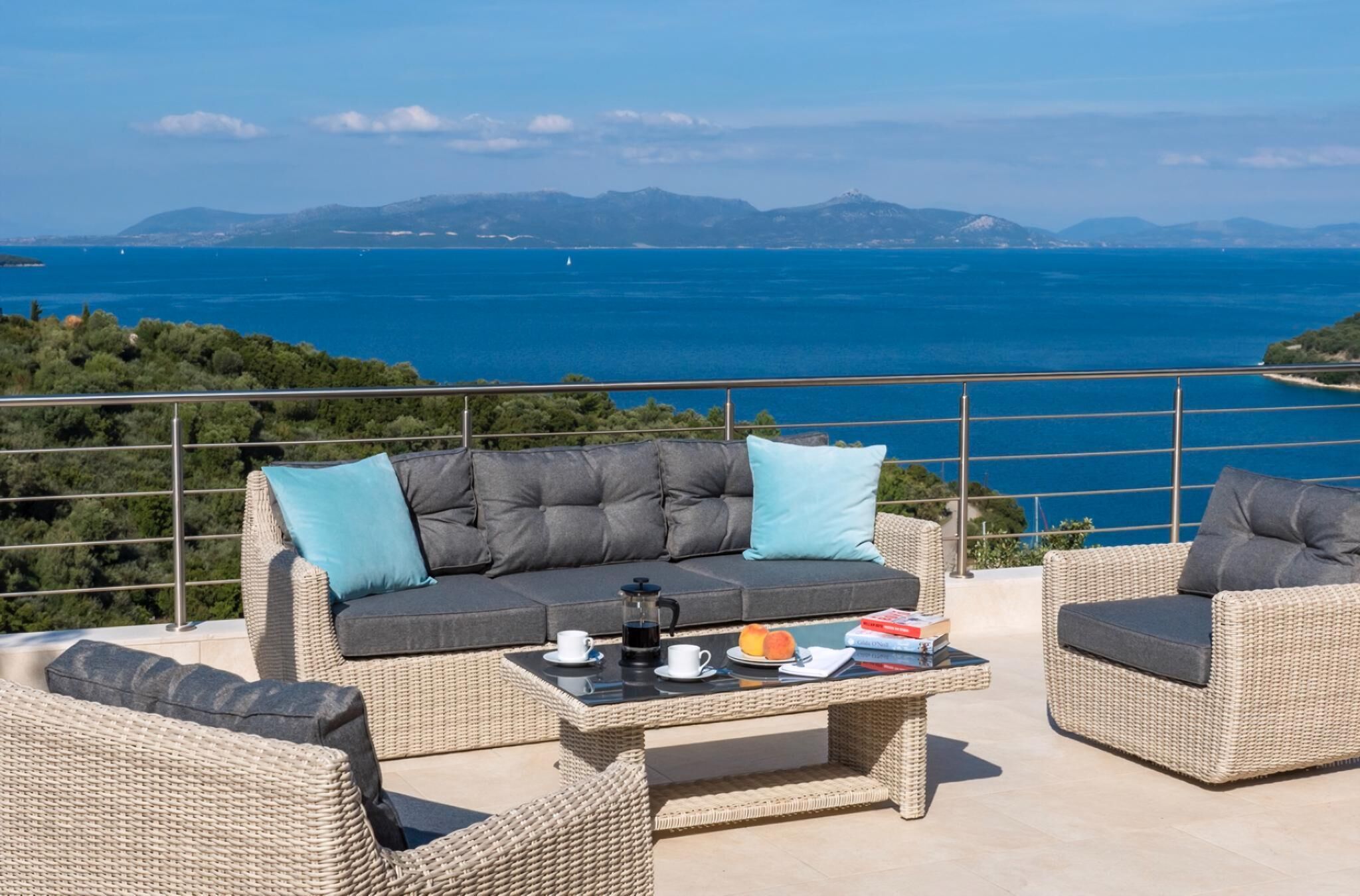 A balcony with wicker furniture, a coffee table, and a view of the sea and mountains.