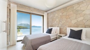 Habitación de hotel con dos camas, pared de piedra y ventana grande con vistas al mar y a las montañas.
