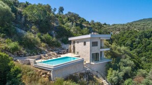 Una villa moderna con piscina, muros de piedra y vistas a las colinas boscosas circundantes.