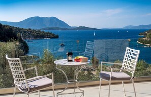 Ein Balkon mit zwei weißen Stühlen und einem kleinen Tisch, mit Blick auf die Küste, wo Boote und Berge zu sehen sind.