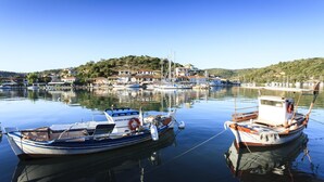 Ein Yachthafen mit mehreren vertäuten Booten, darunter ein kleines Boot mit Kabine, vor der Kulisse von Häusern und Hügeln.