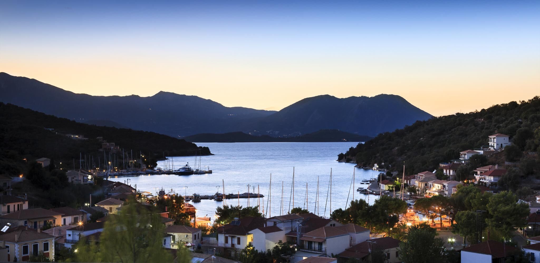 A coastal town at dusk with boats docked at the marina, surrounded by residential buildings and mountains in the background.