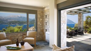 Ein modernes Interieur mit Steinwänden, großen Fenstern mit Blick auf einen See und Berge, sowie eine Terrasse mit Gartenmöbeln.