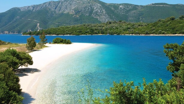Ein Strand mit klarem türkisblauem Wasser, weißem Sand und grünen Hügeln im Hintergrund.
