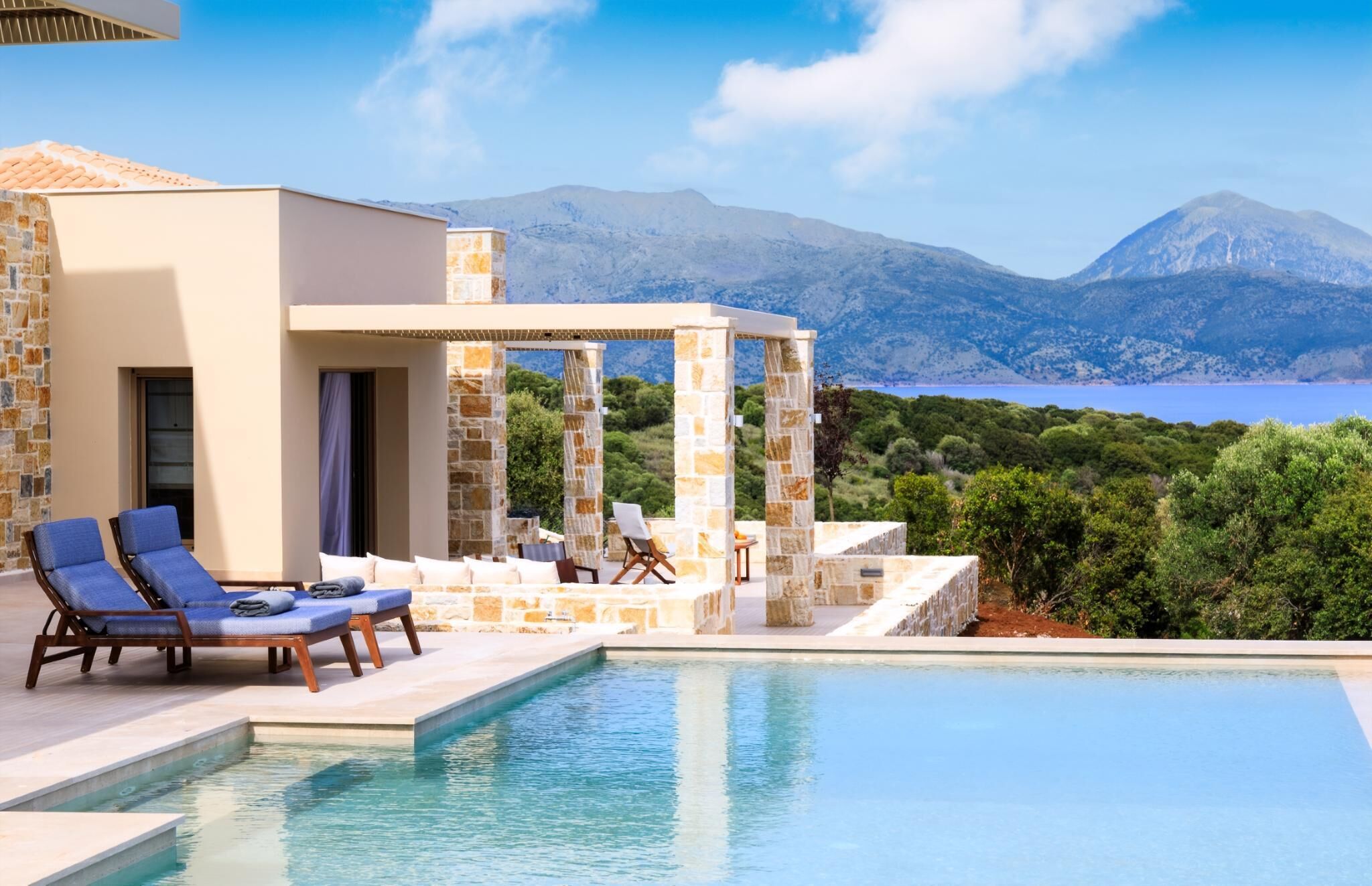 A modern poolside area with lounge chairs, a stone structure, and a view of the sea and mountains.
