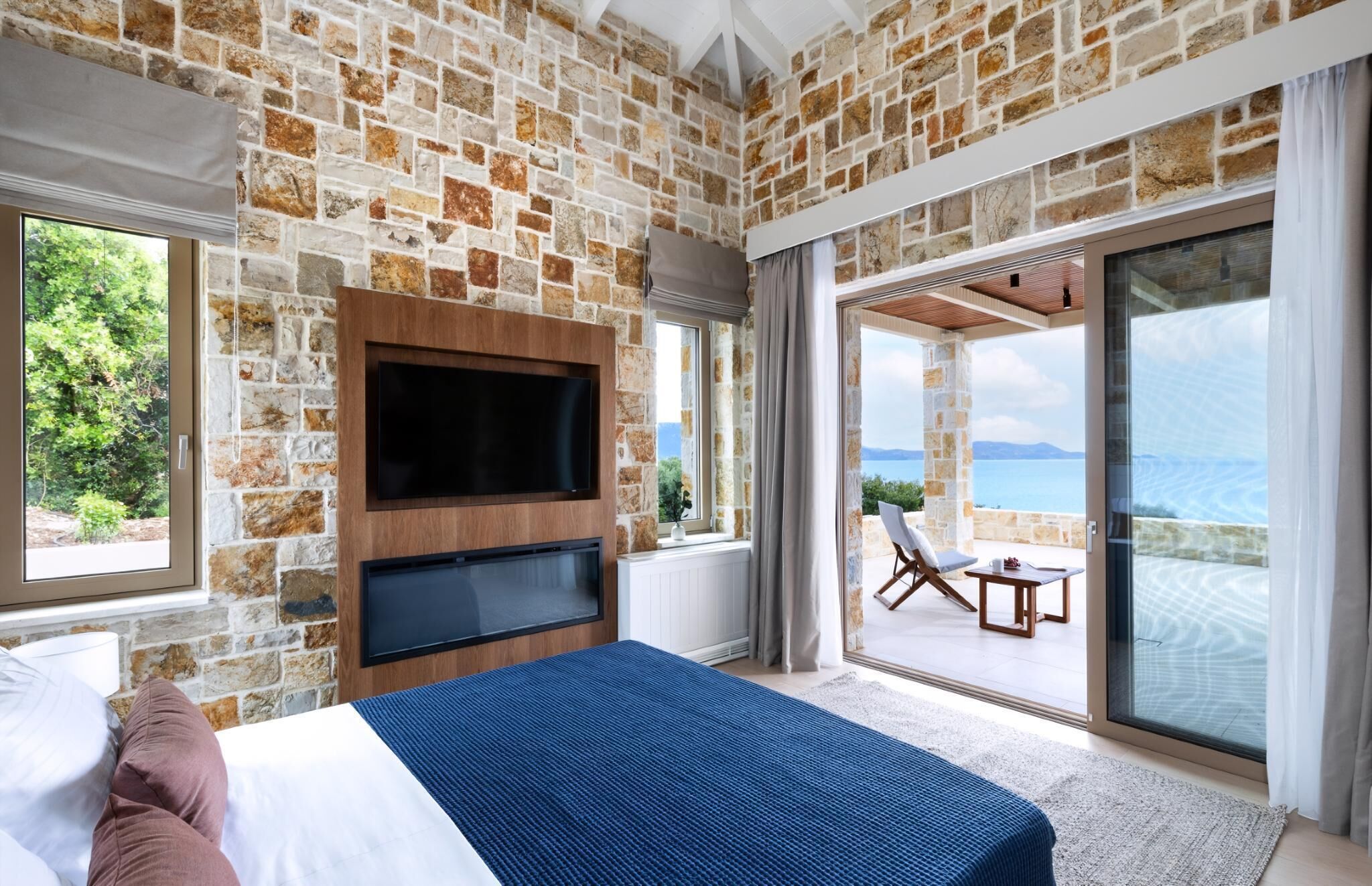 Quarto com parede de pedra, TV de tela plana, cama com colcha azul e varanda com vista para o mar.
