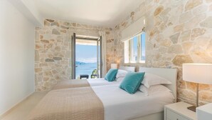 A bedroom with a stone wall, a bed with white bedding and teal pillows, a bedside table with a lamp, and a view of the sea through an open door. - Stylish Contemporary Villa with Sea Views near Vathy Tavernas (Meganissi)