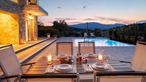 A dining table set for two with a view of a swimming pool and mountains in the background. - Stylish Contemporary Villa with Spacious Terrace near Vathy Tavernas (Meganissi)