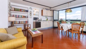 Ein modernes Wohnzimmer mit Sofa, Couchtisch und Essbereich mit Stühlen. Es gibt eine Küche mit Schränken und einem Fenster mit Blick.