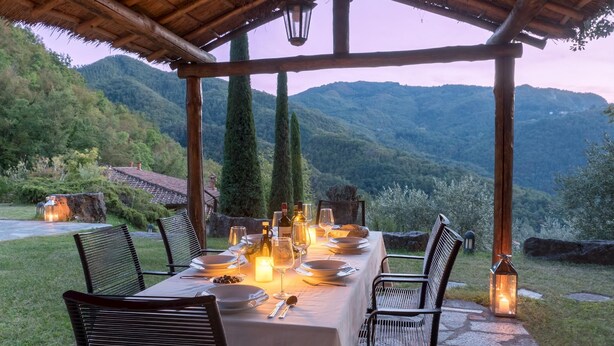Een buitenontbijtplaats met een tafel voor vier personen, omringd door stoelen, onder een houten pergola met uitzicht op een bergachtig landschap.