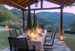 Zona pranzo all'aperto con tavolo imbandito per quattro persone, circondato da sedie, sotto un pergolato di legno con vista su un paesaggio montuoso.