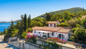 Une maison située sur la côte, avec un toit recouvert de tuiles, entourée d’une végétation luxuriante et offrant une vue sur la mer.