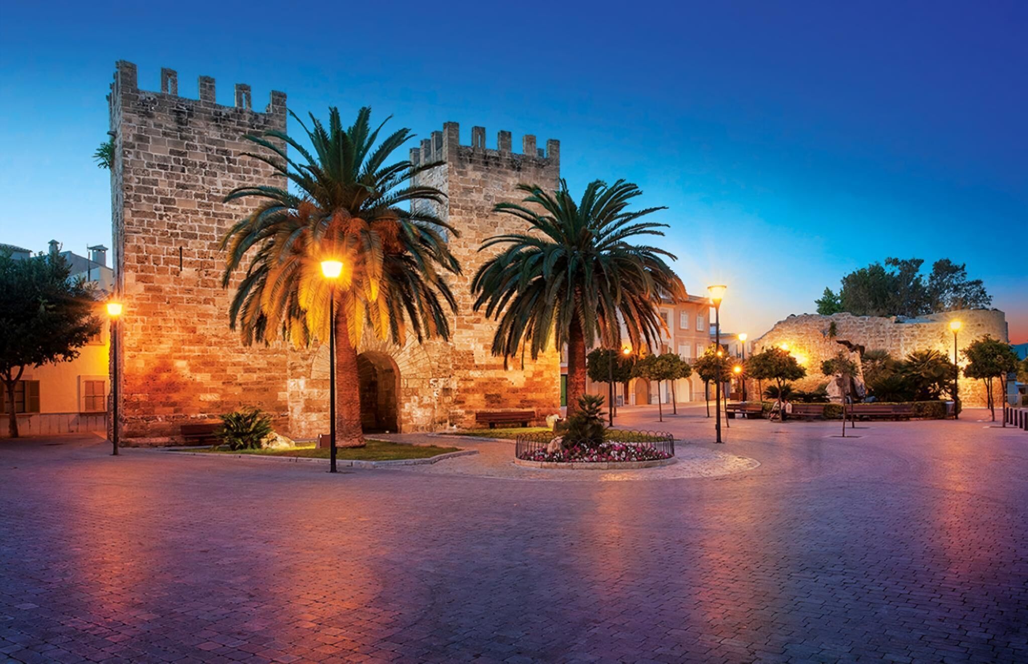 Un edificio histórico de piedra con un amplio arco de entrada, palmeras y una plaza pavimentada con farolas.