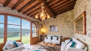 A spacious living room with stone walls, a wooden ceiling, and large windows offering a view of the countryside. - Villa Mille Desideri (Arezzo)