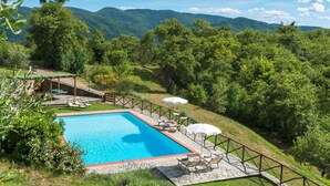 A rectangular swimming pool with a stone deck and a wooden pergola with seating. - Villa Mille Desideri (Arezzo)