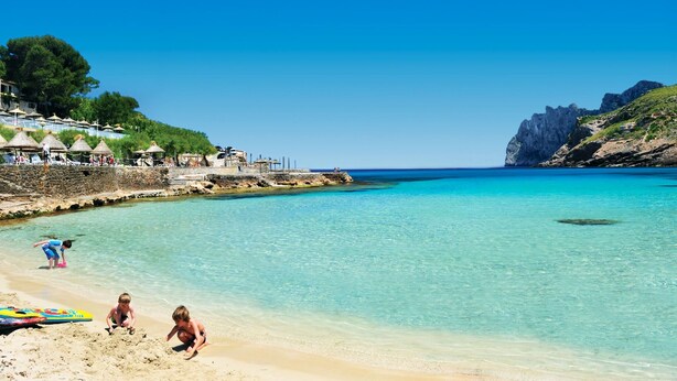 Ein Strand mit klarem türkisblauem Wasser, einem sandigen Ufer und einer felsigen Klippe im Hintergrund.
