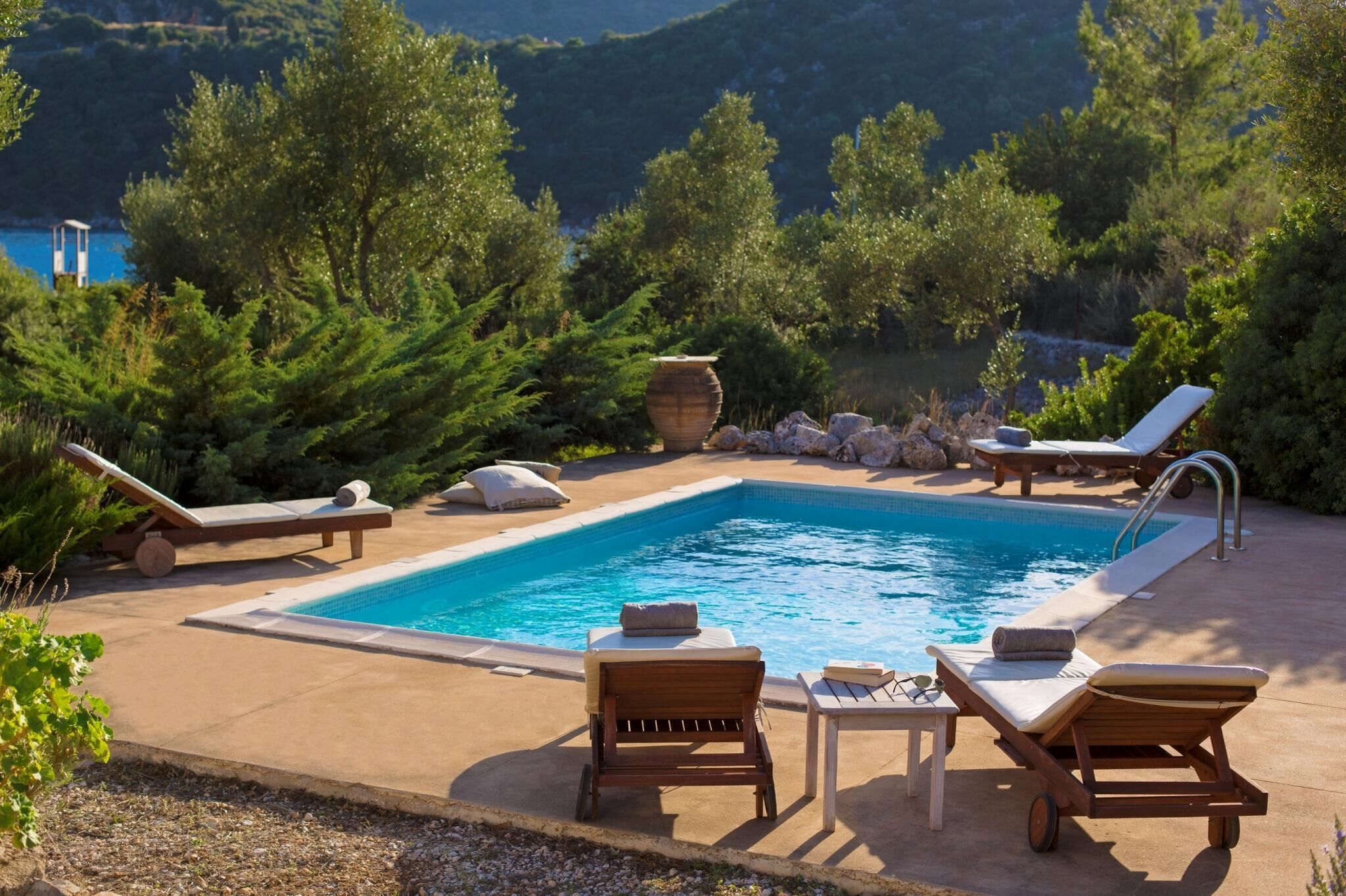 Uma área de piscina com espreguiçadeiras e uma pequena mesa, cercada por vegetação e vista para colinas.
