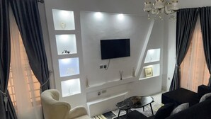 A living room with a flat-screen TV mounted on the wall, a beige armchair, a black sofa, and a small coffee table. - 3 Bedroom En-Suite, Wi-Fi, Secured, 24/7 Power (Lekki)