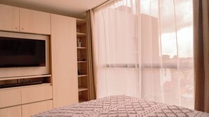 Ein Schlafzimmer mit einem großen Bett, einem an der Wand montierten Fernseher und Blick auf die Stadt durch transparente Vorhänge.