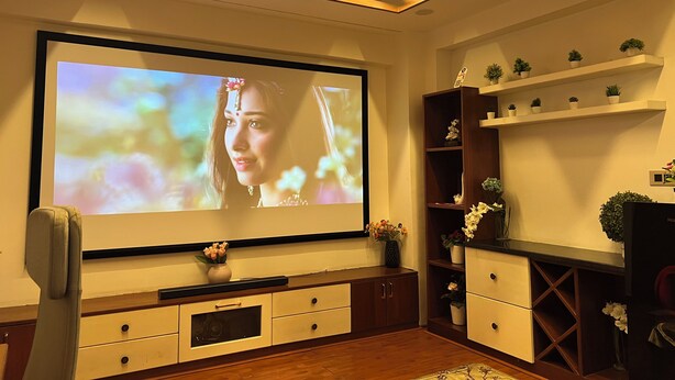 A living room with a large flat-screen TV mounted on the wall, wooden cabinets, a small table with a vase of flowers, and a ceiling fan. - Atlas Homes Penthouse 1BHK Jacuzzi @ Hitech City (Hyderabad)