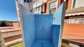 Un espacio de ducha al aire libre con azulejos azules y un desagüe central, rodeado de césped y un edificio con balcones al fondo.